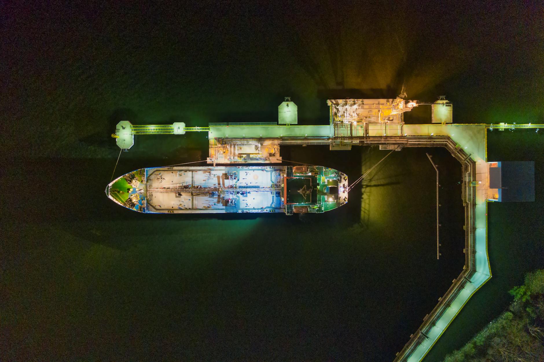An aerial view of a cargo ship docked at a brightly illuminated pier during nighttime.