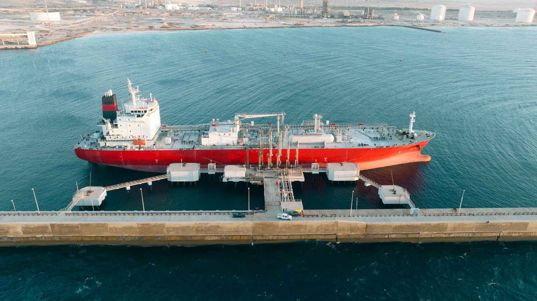 Aerial shot of a red cargo ship docked at an industrial port with clear blue water.